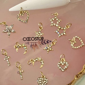 Könnte beinhalten: Sortiment an goldfarbenen Charms mit klaren Strasssteinen. Die Charms umfassen Herzen, Sterne, Monde, Kreuze und eine Blume. Die Charms werden auf einem rosa und goldenen Hintergrund präsentiert.