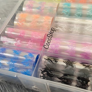 K&ouml;nnte beinhalten: Eine transparente Plastikbox gef&uuml;llt mit Rollen von farbigem Nail-Art-Transferfolie. Die Folie hat verschiedene Flammenmotive in verschiedenen Farben, darunter Pink, Blau, Schwarz und Wei&szlig;. Die Box hat den Text "CocoStop" auf der Seite.
