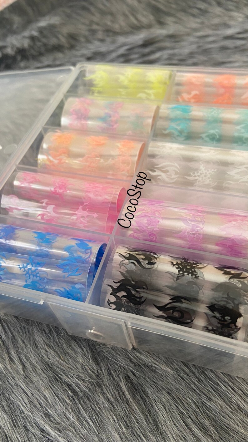 K&ouml;nnte beinhalten: Eine transparente Plastikbox gef&uuml;llt mit Rollen von farbigem Nail-Art-Transferfolie. Die Folie hat verschiedene Flammenmotive in verschiedenen Farben, darunter Pink, Blau, Schwarz und Wei&szlig;. Die Box hat den Text "CocoStop" auf der Seite.