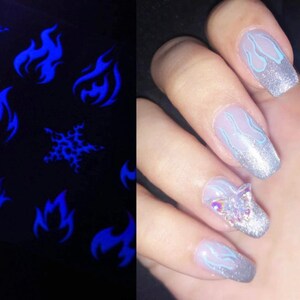 K&ouml;nnte beinhalten: Nahaufnahme einer Hand mit mehreren Fingern&auml;geln, die mit einem silbernen Glitzerlack und einem blauen Flammenmuster lackiert sind. Ein Nagel hat einen klaren Schmetterlings-Strassstein-Akzent.
