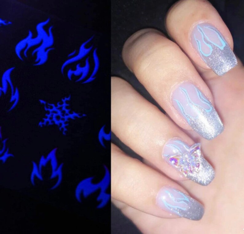 K&ouml;nnte beinhalten: Nahaufnahme einer Hand mit mehreren Fingern&auml;geln, die mit einem silbernen Glitzerlack und einem blauen Flammenmuster lackiert sind. Ein Nagel hat einen klaren Schmetterlings-Strassstein-Akzent.