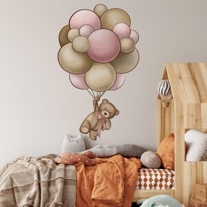 Könnte beinhalten: Ein brauner Teddybär hält an einem Bündel rosa und brauner Ballons fest. Die Ballons schweben über einem Bett mit einer braunen Decke und orange-weiß gemusterter Bettwäsche.