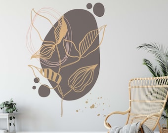 May include: A wall decal featuring a stylized branch with leaves in gold and brown tones. The branch is set against a brown oval shape with pink and white accents. The decal is displayed on a white wall with a wicker chair and a potted plant.
