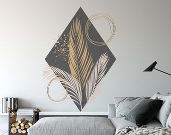 May include: A gray diamond-shaped wall decal with gold and pink palm leaves and gold circles. The decal is on a white wall behind a gray couch with a black throw blanket.