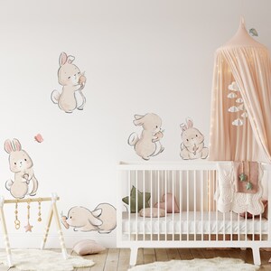 May include: A set of five beige bunny wall decals with pink accents. The bunnies are holding carrots and are arranged around a white crib with a pink and green blanket.