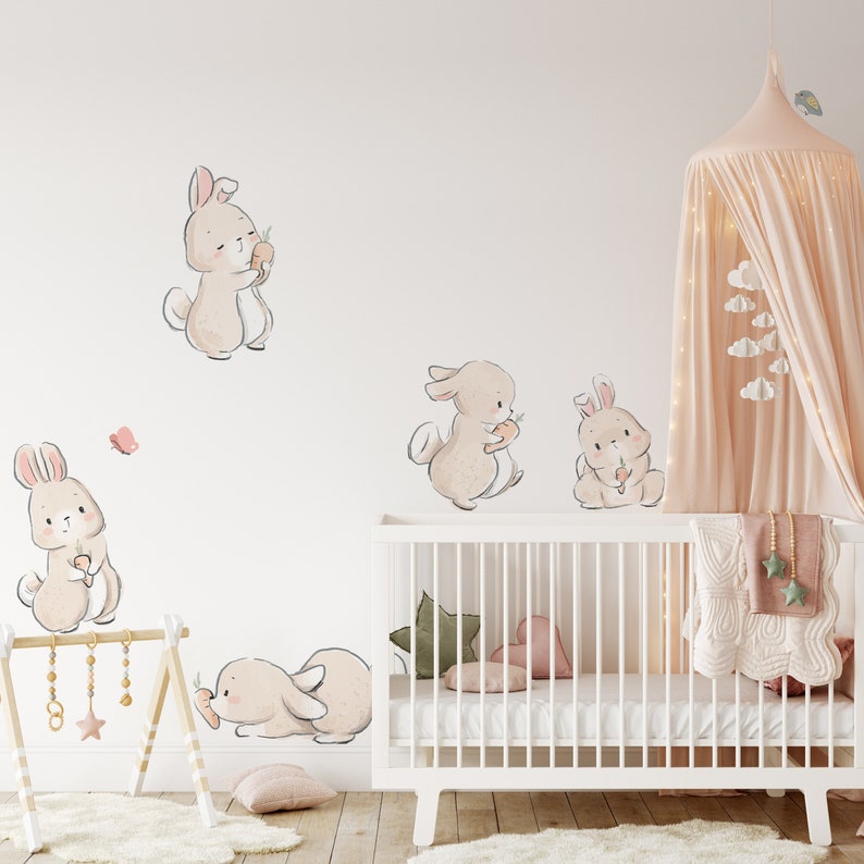 May include: A set of five beige bunny wall decals with pink accents. The bunnies are holding carrots and are arranged around a white crib with a pink and green blanket.