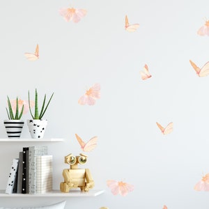 May include: A white wall with a variety of pink and orange butterfly wall decals. The decals are arranged in a random pattern and are of varying sizes. There are two potted plants on a white shelf below the decals.