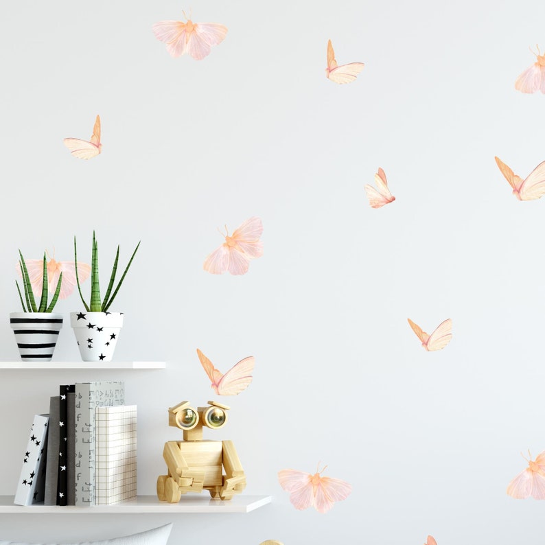 May include: A white wall with a variety of pink and orange butterfly wall decals. The decals are arranged in a random pattern and are of varying sizes. There are two potted plants on a white shelf below the decals.