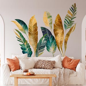 May include: A white couch with pillows and a throw blanket in front of a wall with a large decal of gold and green tropical leaves.
