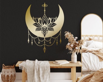 Puede incluir: Una luna creciente dorada con un diseño de flor de loto y cuentas colgantes cuelga de una pared negra. Un banco de madera con una manta se encuentra frente a la pared. Una cesta de mimbre y un jarrón con flores secas están en el suelo.