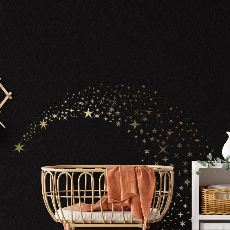 May include: A wicker bassinet with a peach-coloured blanket against a black wall featuring a gold star design. The stars are arranged in a curved line, creating a shooting star effect.