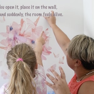 May include: A wall decal featuring a cluster of pink and purple butterflies. The text "You open it, place it on the wall, and suddenly, the room feels alive" is printed above the butterflies. Two people are shown applying the decal to a white wall.