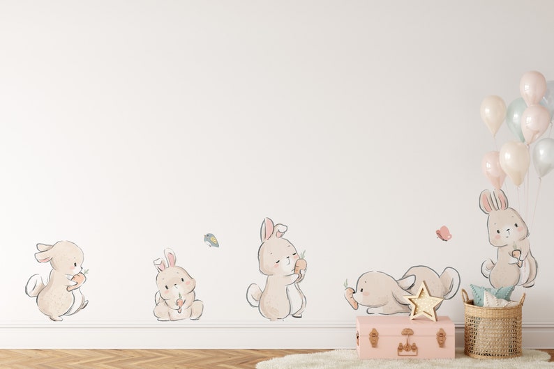 May include: A white wall with five brown bunny rabbit wall decals. The bunnies are holding carrots and there are butterflies on the wall. There is a pink suitcase and a wicker basket with a light blue and white blanket in the bottom right corner of the image. There are pastel pink, blue, and white balloons in the top right corner of the image.