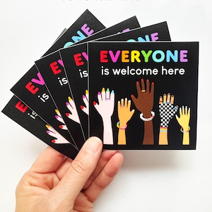 May include: A hand holds a stack of black cards with the text "EVERYONE is welcome here" in rainbow colors. The cards feature illustrations of hands with diverse skin tones and accessories. The top of the image reads "100% OF PROFITS GO TO THE ACLU."