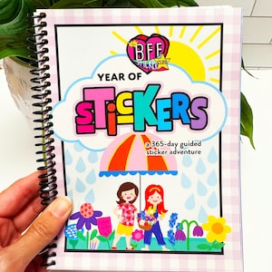 May include: A spiral-bound book titled Year of Stickers, a 365-day guided sticker adventure. The cover features a colorful illustration of two figures under a striped umbrella, surrounded by flowers and rain, with a BFF Sticker Club logo.
