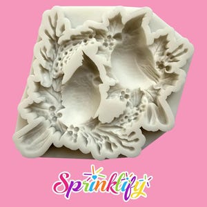 May include: A white silicone mold featuring a bird design surrounded by holly leaves and berries. The mold is square-shaped with detailed relief patterns, perfect for crafting. The word "Sprinklify" is written in a colorful, cursive font at the bottom.