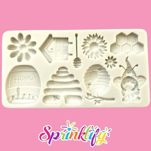 Könnte beinhalten: Eine weiße Silikonform mit verschiedenen Bienenmotiven. Enthält eine Blume, einen Bienenstock, einen Honiglöffel, eine Honigwabe und einen Bienenzwerg. Das Wort "HONSA" ist auf einer fassförmigen Form zu sehen. Die Form befindet sich auf einem rosa Hintergrund.