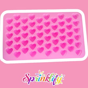 Peut inclure: Un moule en silicone rose avec un bord blanc, rempli de 48 cavités en forme de cœur. Le mot "Sprinklify" est écrit en bas dans une police cursive colorée. Ce moule est idéal pour faire des bonbons, des chocolats ou d'autres friandises.