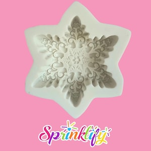 May include: A white silicone snowflake mold with intricate floral and scrollwork details. The mold is in the shape of a six-pointed star, perfect for creating decorative elements for baking or crafts. The background is a solid pink.