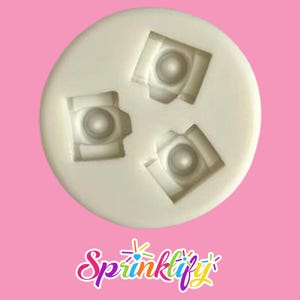 May include: A white silicone mold with three camera-shaped cavities. Each cavity features a detailed camera design with a central lens and rectangular frame. The mold is circular and sits against a pink background. The word "Sprinklify" is written in a colorful script.