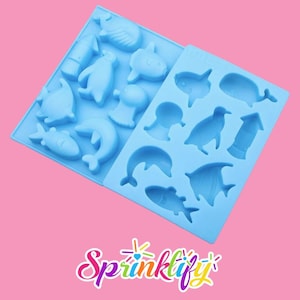 May include: Two light blue silicone molds with various sea creature shapes, including fish, dolphins, penguins, and whales. The molds are on a pink background, with the word "Sprinklify" in a cursive font at the bottom.