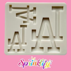 May include: Off-white silicone mold featuring the letters "LA" in a stacked arrangement. The mold is designed for creating decorative elements. The background is a solid pink color, and the logo "Sprinklify" is at the bottom.
