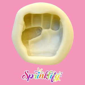 Puede incluir: Un molde de silicona amarillo claro con forma de puño cerrado. El molde tiene impresiones detalladas de los dedos y la palma. El fondo es de color rosa sólido y el logotipo "Sprinklify" está en la parte inferior.