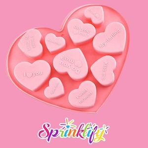 Puede incluir: Un molde rosa en forma de corazón con múltiples cavidades en forma de corazón. Cada corazón tiene una frase romántica grabada, como "Te amo" y "Bésame". La palabra "Sprinklify" está en la parte inferior.