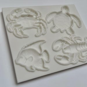 May include: A white rectangular silicone mold featuring four recessed sea creature designs: a crab, a sea turtle, an angelfish, and a lobster. The mold has a smooth surface with detailed indentations for each marine animal shape.