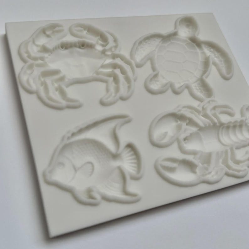 May include: A white rectangular silicone mold featuring four recessed sea creature designs: a crab, a sea turtle, an angelfish, and a lobster. The mold has a smooth surface with detailed indentations for each marine animal shape.