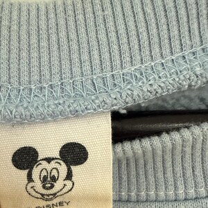 May include: A light blue ribbed crewneck collar on a sweatshirt with an off-white fabric tag attached. The tag features a black line art illustration of Mickey Mouse's face and the text "&copy; DISNEY" printed in black ink below the character's head.