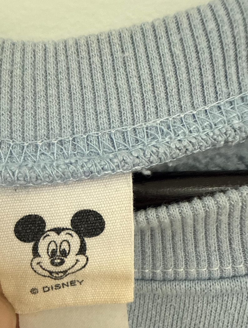 May include: A light blue ribbed crewneck collar on a sweatshirt with an off-white fabric tag attached. The tag features a black line art illustration of Mickey Mouse's face and the text "&copy; DISNEY" printed in black ink below the character's head.