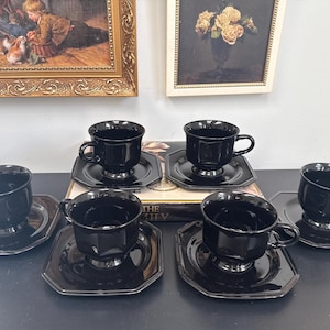 May include: A set of six black teacups and saucers. The cups have a classic design with a square base and a rounded top. The saucers are octagonal. The cups and saucers are displayed on a dark surface. Framed art hangs in the background.