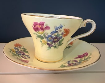 May include: A yellow teacup and saucer set with a floral pattern. The cup and saucer have a gold trim around the edges. The flowers are pink, purple, blue, and yellow.