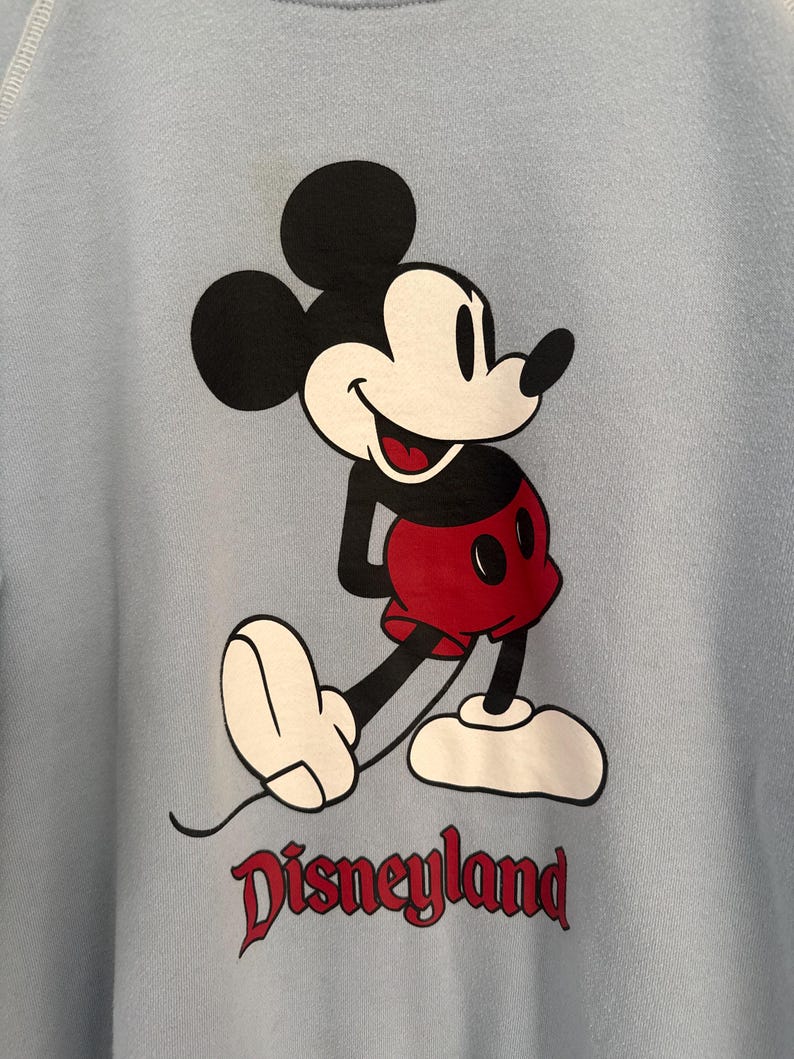 May include: A light blue sweatshirt featuring a graphic of Mickey Mouse in black, white, and red colors. Mickey is depicted in a classic pose with red shorts and white shoes. Below the character, the word Disneyland is printed in a dark red font.