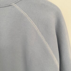 May include: A light blue sweatshirt featuring a ribbed crew neckline and raglan sleeves. The garment is constructed with prominent white topstitching along the diagonal seam that connects the sleeve to the main body of the pullover.