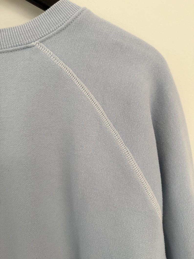 May include: A light blue sweatshirt featuring a ribbed crew neckline and raglan sleeves. The garment is constructed with prominent white topstitching along the diagonal seam that connects the sleeve to the main body of the pullover.