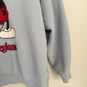 May include: A light blue long-sleeve sweatshirt featuring a graphic of Mickey Mouse wearing red shorts and white shoes. The text Disneyland is printed in red below the character. The garment has a ribbed crew neckline, cuffs, and hem.