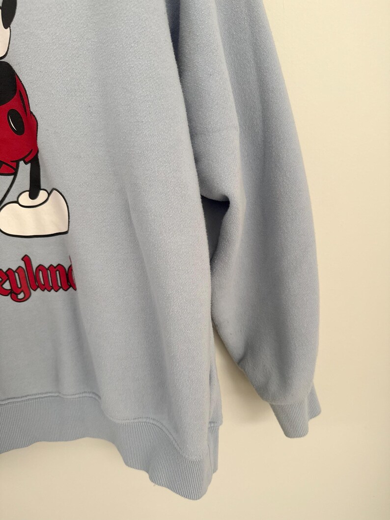 May include: A light blue long-sleeve sweatshirt featuring a graphic of Mickey Mouse wearing red shorts and white shoes. The text Disneyland is printed in red below the character. The garment has a ribbed crew neckline, cuffs, and hem.