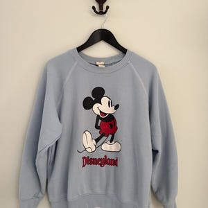 May include: A light blue crewneck sweatshirt featuring a graphic of Mickey Mouse in red shorts and white shoes. The word Disneyland is printed in red lettering below the character. The garment has long sleeves with ribbed cuffs and a ribbed neckline.