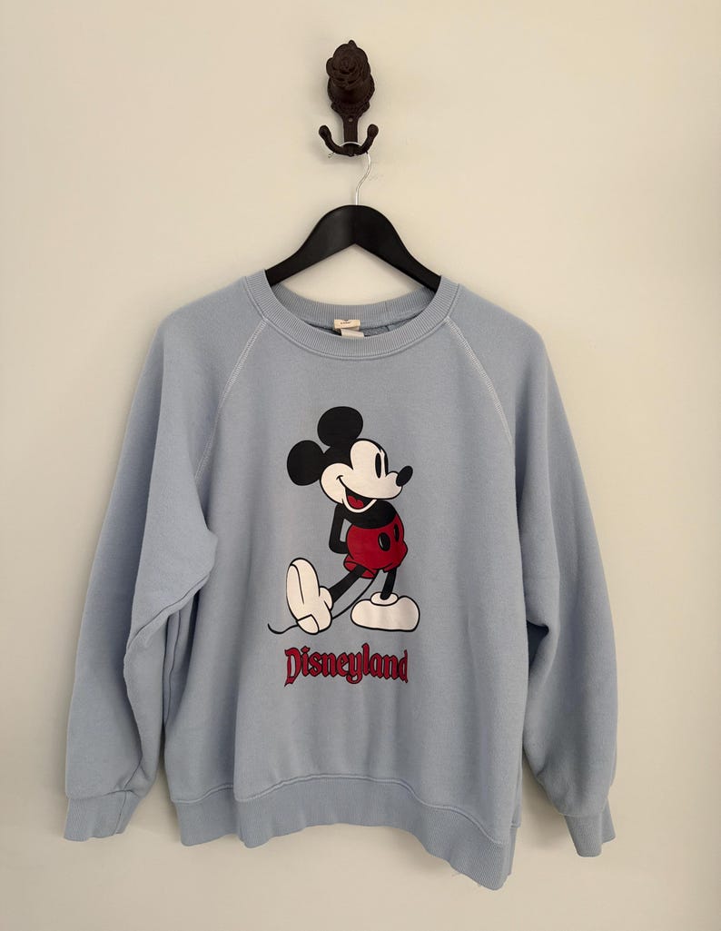 May include: A light blue crewneck sweatshirt featuring a graphic of Mickey Mouse in red shorts and white shoes. The word Disneyland is printed in red lettering below the character. The garment has long sleeves with ribbed cuffs and a ribbed neckline.