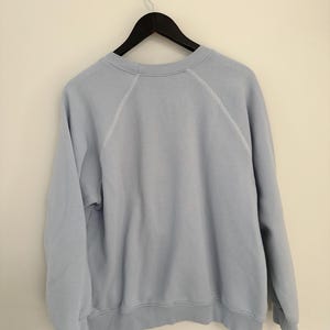 May include: A light blue crewneck sweatshirt featuring long raglan sleeves with visible white stitching along the shoulder seams. The garment has a ribbed collar, ribbed cuffs, and a ribbed hem, hanging on a black hanger against a plain wall.