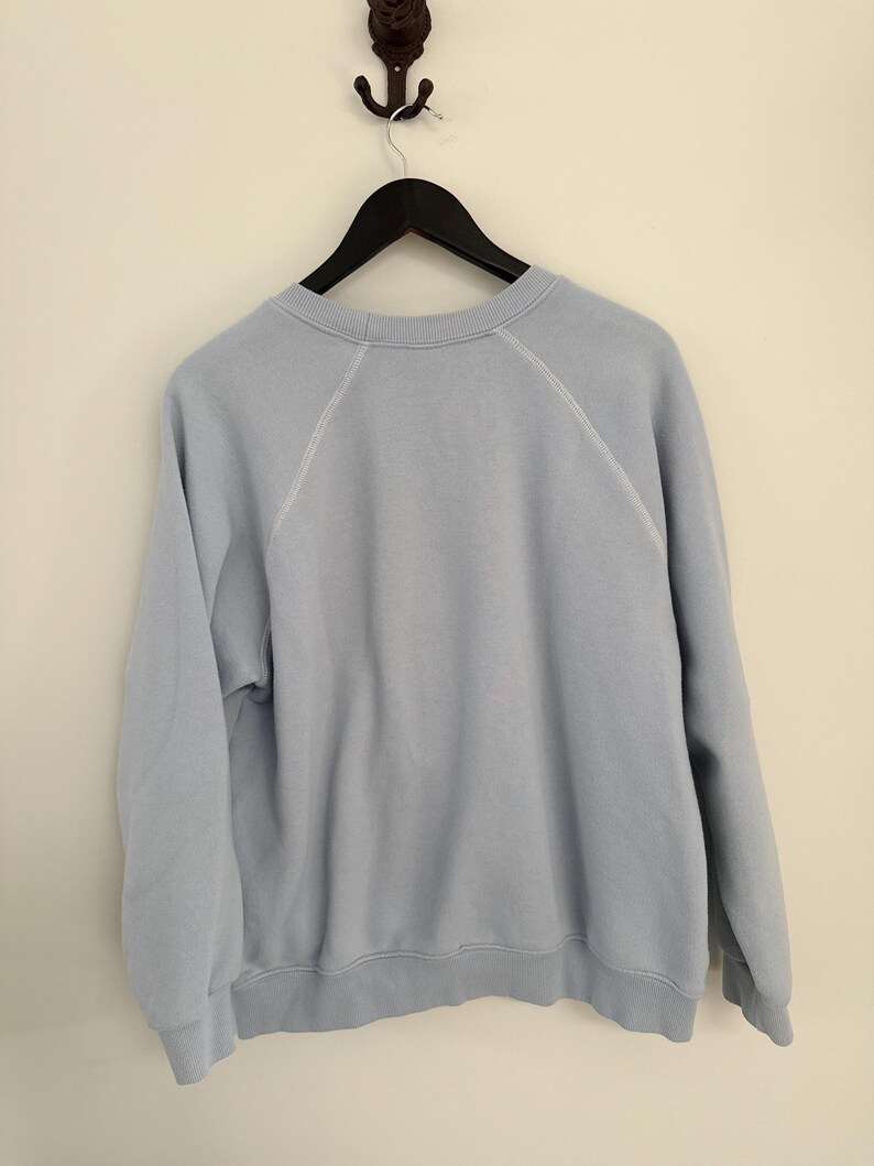 May include: A light blue crewneck sweatshirt featuring long raglan sleeves with visible white stitching along the shoulder seams. The garment has a ribbed collar, ribbed cuffs, and a ribbed hem, hanging on a black hanger against a plain wall.
