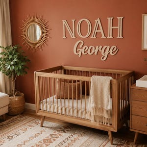 K&ouml;nnte beinhalten: Ein h&ouml;lzernes Kinderbett mit vertikalen St&auml;ben, einer hellen Matratze und einer Decke mit Fransen. &Uuml;ber dem Bett sind gro&szlig;e Holzbuchstaben, die den Namen NOAH George in einer Kombination aus Block- und Schreibschrift bilden, an der Wand befestigt.