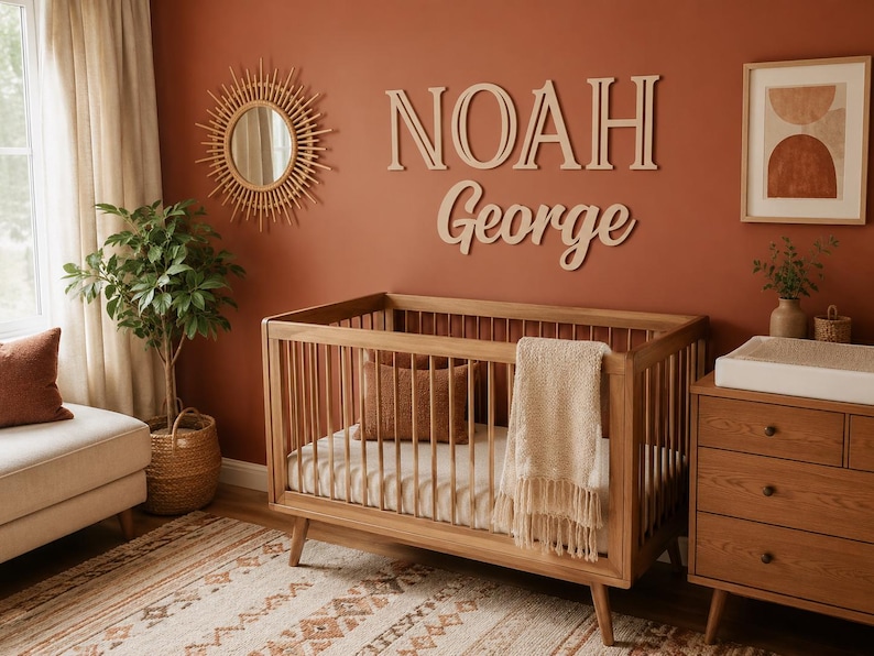 K&ouml;nnte beinhalten: Ein h&ouml;lzernes Kinderbett mit vertikalen St&auml;ben, einer hellen Matratze und einer Decke mit Fransen. &Uuml;ber dem Bett sind gro&szlig;e Holzbuchstaben, die den Namen NOAH George in einer Kombination aus Block- und Schreibschrift bilden, an der Wand befestigt.