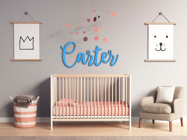 May include: A light blue wooden name sign spelling "Carter" hangs above a crib in a nursery. The crib has a white frame and a pink mattress. There are two framed prints on the wall, one with a crown and the other with a cartoon animal face.