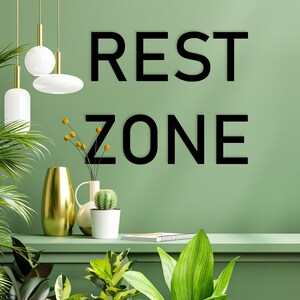 May include: A green wall with the text "REST ZONE" in black lettering. There are two hanging lights, a gold vase, a white vase with a cactus, and a book on a shelf.