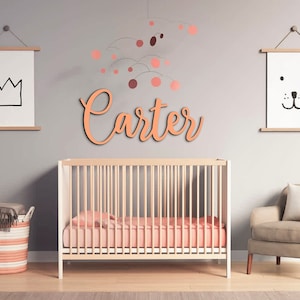 May include: A nursery room with a crib, a chair, and a basket. The walls are painted a light gray color. There is a wooden name sign that says "Carter" above the crib. There is a mobile hanging from the ceiling above the crib.