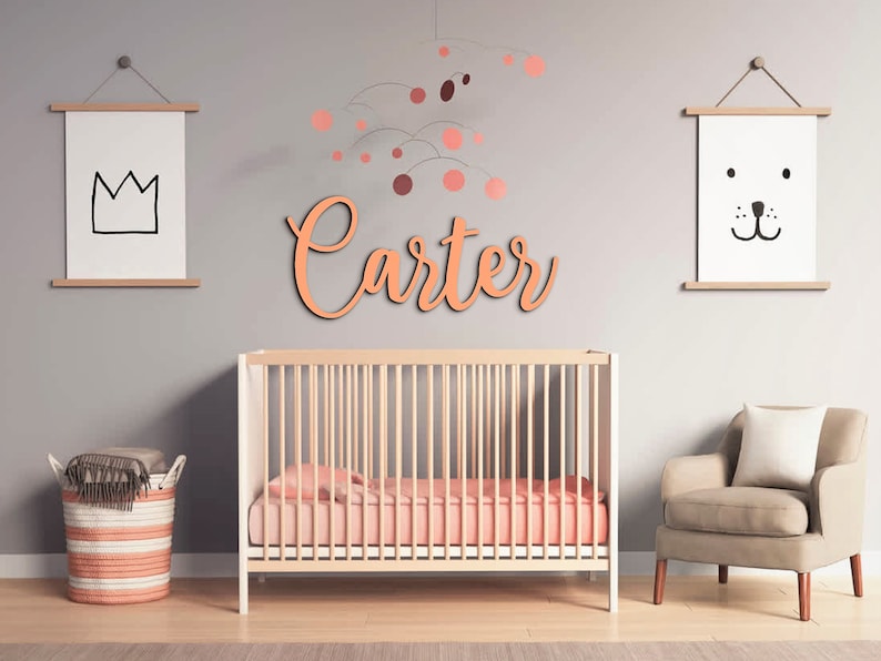 May include: A nursery room with a crib, a chair, and a basket. The walls are painted a light gray color. There is a wooden name sign that says "Carter" above the crib. There is a mobile hanging from the ceiling above the crib.