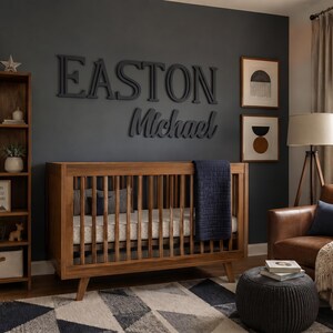 K&ouml;nnte beinhalten: Ein h&ouml;lzernes Kinderbett mit vertikalen St&auml;ben und einer dunkelblauen Decke, platziert in einem Raum mit einem hohen Holzregal, einem braunen Ledersessel, einer dunkelgrauen Wand mit dem Namen EASTON Michael in gro&szlig;en Buchstaben und einem geometrischen Teppich.